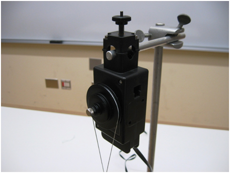 A clamp on the top of a ring stand holds a horizontal rod.  An apparatus is attached to the end of the rod with a thumbscrew.  The bulk of the apparatus is below the rod.  On the side of this apparatus are two vertical, concentric pullies of different radii, but that are all attached to each other.  A cord passes around the largest pulley and the cords appear to cross just below the bottom edge of the picture.